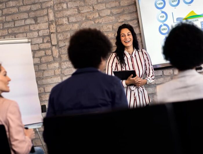 a group of employees during a meeting with a trainer at the head of the room with a clipboard.