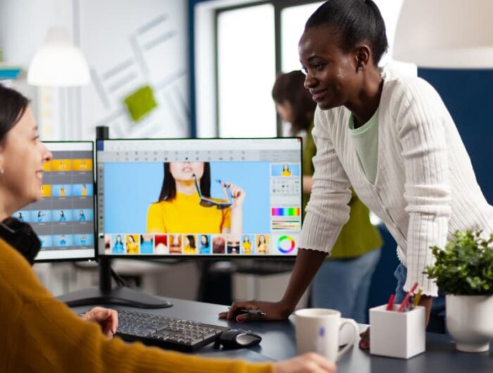 Two female coworkers talking at a desk next to a computer with the newest computer software.