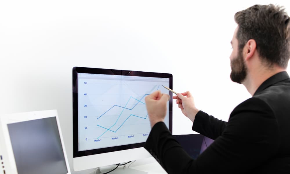 A businessman sits at a laptop computer showing a chart with a pencil in one hand. He’s making a fist of victory with the other.
