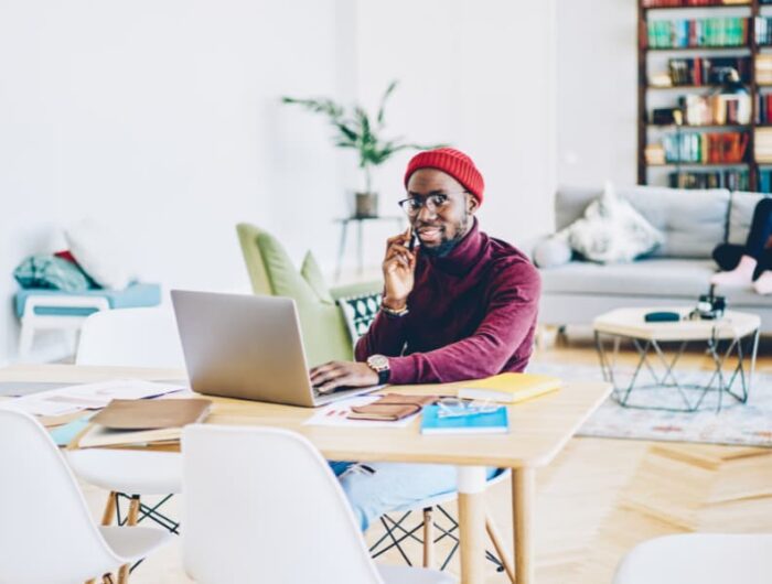 A black man working remotely on a laptop and talking on a smartphone.