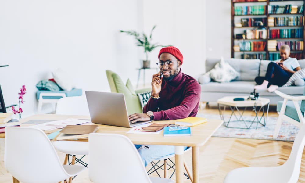 A black man working remotely on a laptop and talking on a smartphone.